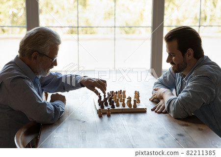Side view young man with mature father playing chess together Side view young man with mature father playing chess together 82211385