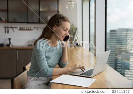 Young Caucasian woman talk on smartphone working on computer Young Caucasian woman talk on smartphone working on computer 82211502