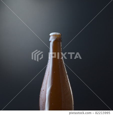 Front view of a bottle of cold beer with overflowing foam against dark blue grunge background. Cold alcoholic beverage, international beer day concept. Organic photography style. Front view of a bottle of cold beer with overflowing foam against dark blue grunge background. Cold alcoholic beverage, international beer day concept. Organic photography style. 82213995