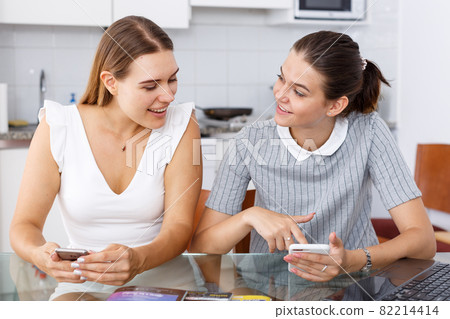 Young smiling girlfriends using phones and chatting table Young smiling girlfriends using phones and chatting table 82214414