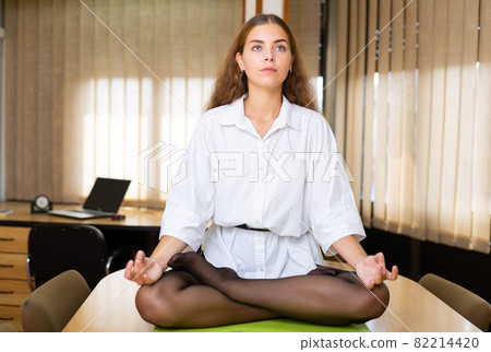 Relaxed young woman sitting in lotus position on office desk 82214420
