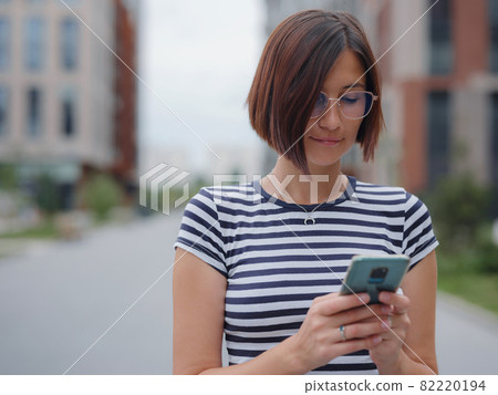 Smiling woman using mobile phone in the street Smiling woman using mobile phone in the street 82220194