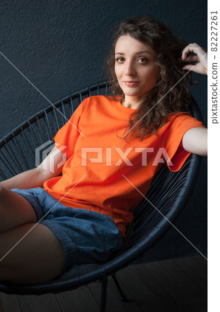 Female portrait of a beautiful curly girl in bright orange t-shirt sitting at home in her apartment on dark background, happy people concept 82227261