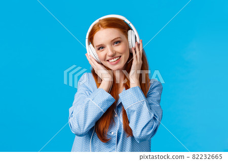 Technology, lifestyle and people concept. Cute redhead woman in pyjama, listen music in headphones, enjoy nice beats in earphones, good quality, new songs, standing blue background 82232665