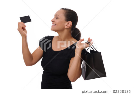 Portrait of young mixed race African American woman smiling with beautiful toothy smile holding shopping bags and credit or discount card in hands, standing against white background with copy space Portrait of young mixed race African American woman smiling with beautiful toothy smile holding shopping bags and credit or discount card in hands, standing against white background with copy space 82238185
