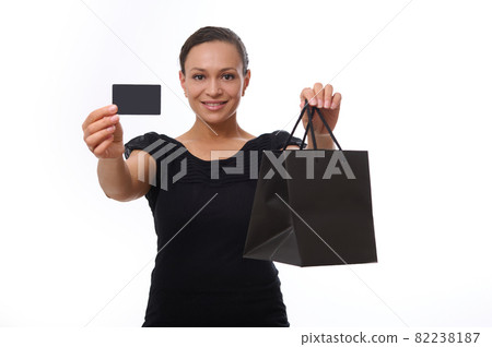 Beautiful mixed race woman looks at camera and smiles toothy smile, showing black shopping packet and plastic credit or discount card in her outstretched hands. Concept of Black Friday with copy space Beautiful mixed race woman looks at camera and smiles toothy smile, showing black shopping packet and plastic credit or discount card in her outstretched hands. Concept of Black Friday with copy space 82238187