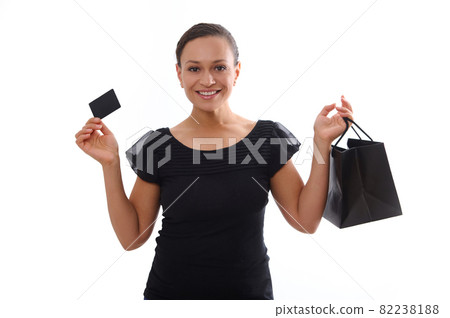Isolated portrait on white background with copy space of attractive smiling woman dressed in black and holding a discount credit card and a shopping bag, looking at camera. Black Friday concept Isolated portrait on white background with copy space of attractive smiling woman dressed in black and holding a discount credit card and a shopping bag, looking at camera. Black Friday concept 82238188