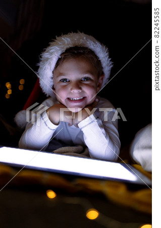 Happy caucasian boy wearing santa hat, using tablet, looking at camera at christmas time 82245585