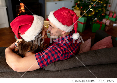 Happy caucasian couple wearing santa hats and embracing on sofa at christmas time Happy caucasian couple wearing santa hats and embracing on sofa at christmas time 82245629