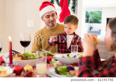 Happy caucasian father and son wearing santa hats sitting at christmas table Happy caucasian father and son wearing santa hats sitting at christmas table 82245798