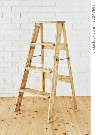 Antique wooden stepladder placed in front of a white brick wall 82247692
