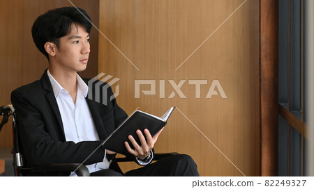 Disabled businessman in wheelchair holding notebook and looking out of window while sitting in office. 82249327