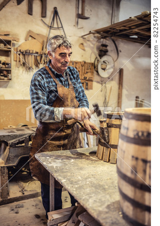 Carpenter places metal stripe on a new wooden barrel made in his cozy workshop 82254743