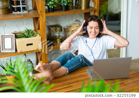 Smiling woman in headset in front of a laptop monitor at home. She puts her feet on the table and vividly discusses the latest gossip with her friend. Smiling woman in headset in front of a laptop monitor at home. She puts her feet on the table and vividly discusses the latest gossip with her friend. 82255199