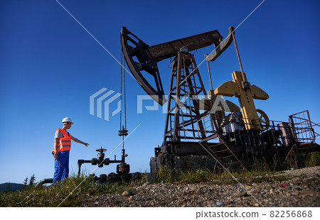 Male worker in work vest holding industrial wrench and pointing finger at petroleum pump jack while working in oil field. Petroleum engineer standing near oil pump rocker-machine under blue sky. Male worker in work vest holding industrial wrench and pointing finger at petroleum pump jack while working in oil field. Petroleum engineer standing near oil pump rocker-machine under blue sky. 82256868