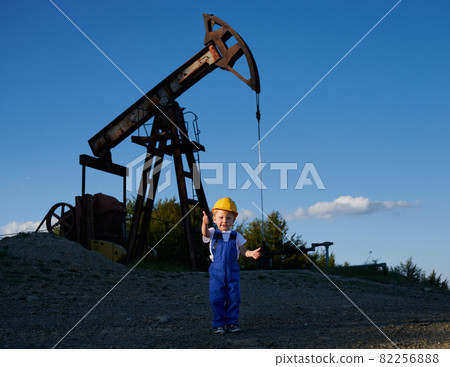 Young child boy in working clothing and helmet with happy face expression standing on stone surface and showing thumb up. Behind him oil pumping machinery in operation under blue sky. Young child boy in working clothing and helmet with happy face expression standing on stone surface and showing thumb up. Behind him oil pumping machinery in operation under blue sky. 82256888