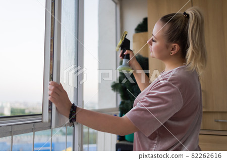 Beautiful young housewife in working clothes washing windows by professional tools at home. 82262616