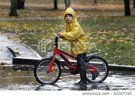 Boy in yellow raincoat on bicycle is riding on puddles. Rainy autumn day 82267290