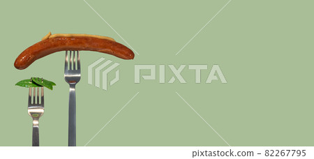Banner with green fresh leaves picked by a fork for eating as new vegan food and grilled sausage with mustard on another fork at salad color solid background with copy space for text, closeup, details Banner with green fresh leaves picked by a fork for eating as new vegan food and grilled sausage with mustard on another fork at salad color solid background with copy space for text, closeup, details 82267795