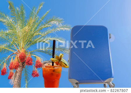 Banner concept with colorful date palm tree with fruits, tropical cocktail and big blue suitcase trolley at blue gradient background with copy space and tropical hot sun lens flare effect 82267807