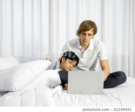 Father work at home with son. A boy napping on his father's lap while the father using laptop computer to work in the bedroom. Work life balance. 82289491