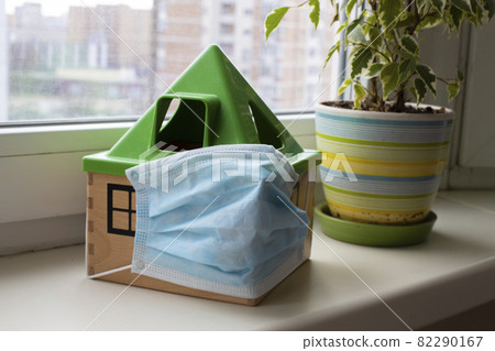 concept obey quarantine coronavirus mode. a toy house in a medical mask stands on a windowsill next to a flower in a pot concept obey quarantine coronavirus mode. a toy house in a medical mask stands on a windowsill next to a flower in a pot 82290167