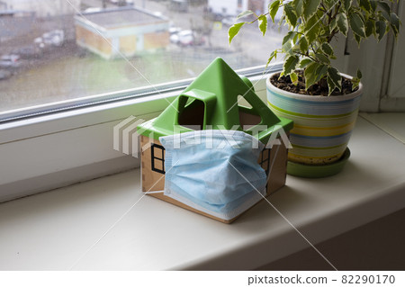 safe home concept, quarantine coronavirus. a toy house in a medical mask stands on a windowsill next to a flower in a pot safe home concept, quarantine coronavirus. a toy house in a medical mask stands on a windowsill next to a flower in a pot 82290170