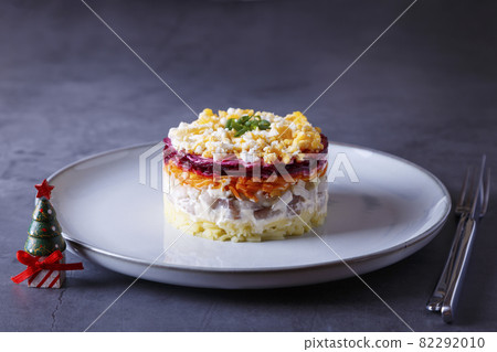 Herring salad under a fur coat. Traditional Russian multilayered salad from herring, beets, potatoes, carrots and eggs. Close-up, grey background, miniature Christmas tree. 82292010