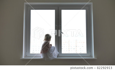 Lonely child girl sitting on the windowsill at the window in the apartmen. Lonely child girl sitting on the windowsill at the window in the apartmen. 82292913