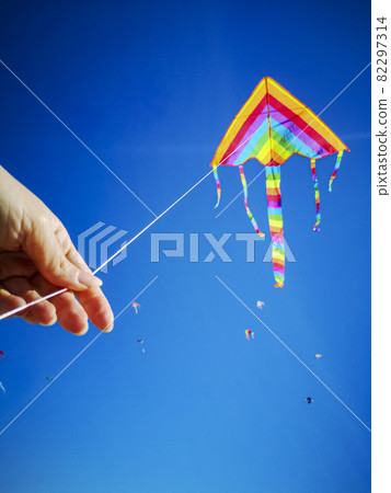 Hand holding multicolored kite in LGBT like colors attached to a white string against blue sky and other kites at festival in Latvia 82297314