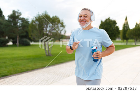 Happy mature man wearing headphones with water bottle in hand jogging outside in city park Happy mature man wearing headphones with water bottle in hand jogging outside in city park 82297618