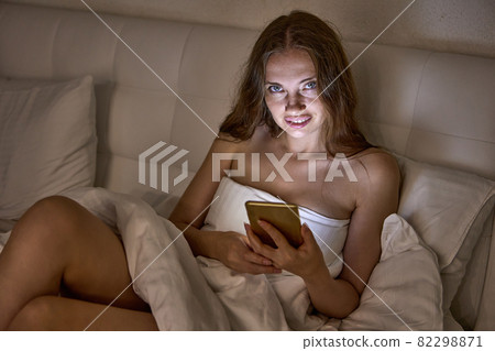 Cheerful bare woman lies in bed with phone. 82298871