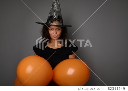 Charming mysterious mixed race woman dressed in black witch carnival Halloween costume and wizard hat, holds orange colorful air balloons, looks at camera, isolated over gray background, copy space. 82311249