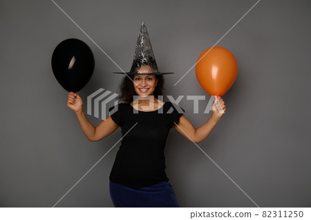 Beautiful attractive woman dressed in witch black Halloween costume and wizard hat, stands against a gray background with orange and black air balloon, smiles looking at camera 82311250