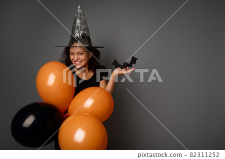 Beautiful mysterious smiling woman in wizard hat, dressed in black, holds orange black air balloons and handmade felt-cut black bat, looks at camera, poses against gray background, copy space . 82311252