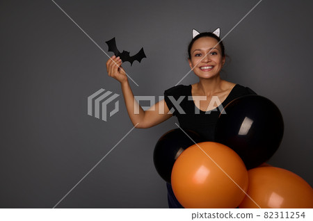 Joyful Hispanic woman dressed in Halloween carnival costume looks at camera, holds handmade bat in her hand , posing with colorful black orange air balloons against gray background with copy space Joyful Hispanic woman dressed in Halloween carnival costume looks at camera, holds handmade bat in her hand , posing with colorful black orange air balloons against gray background with copy space 82311254