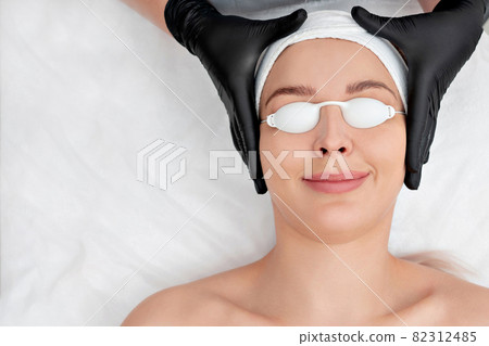 Beautiful young woman with covered eyes is getting face skin treatment. Doctor in medical gloves is going to do the procedure 82312485
