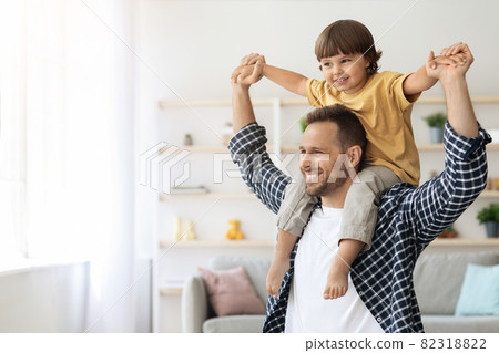 Father's day. Happy little boy sitting on daddy's shoulders and smiling, both looking aside at empty space at home Father's day. Happy little boy sitting on daddy's shoulders and smiling, both looking aside at empty space at home 82318822