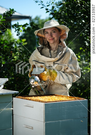 Portrait of a beautiful young female beekeeper working in an apiary near beehives with bees. Collect honey. Beekeeper on apiary. Beekeeping concept. Portrait of a beautiful young female beekeeper working in an apiary near beehives with bees. Collect honey. Beekeeper on apiary. Beekeeping concept. 82318987
