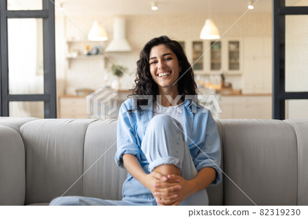 Portrait of pretty young brunette woman smiling and looking at camera, sitting on couhc at home Portrait of pretty young brunette woman smiling and looking at camera, sitting on couhc at home 82319230