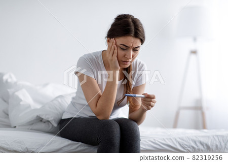 Depressed sad unhappy young pretty european woman looking at negative home test sitting on bed in bedroom interior alone Depressed sad unhappy young pretty european woman looking at negative home test sitting on bed in bedroom interior alone 82319256