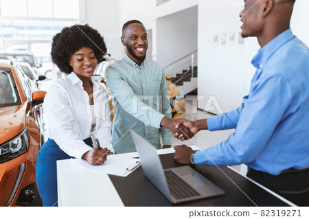 Happy black couple buying new car, shaking hands with seller, signing auto purchase agreement at dealership 82319271