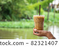 Glass of cold espresso coffee in hand Background blurry views tree and water 82321407