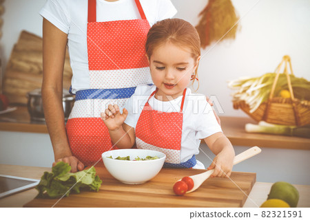 Happy woman and her daughter making healthy vegan salad and snacks for family feasting in sunny kitchen. Christmas, New year, Thanksgiving, Anniversary, Mothers Day. Healthy meal cooking concept Happy woman and her daughter making healthy vegan salad and snacks for family feasting in sunny kitchen. Christmas, New year, Thanksgiving, Anniversary, Mothers Day. Healthy meal cooking concept 82321591
