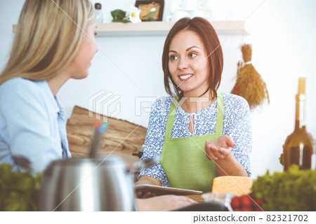 Two women friends choose the recipe and ingredients for a delicious meal while sitting in sunny kitchen. Vegetarian concept Two women friends choose the recipe and ingredients for a delicious meal while sitting in sunny kitchen. Vegetarian concept 82321702
