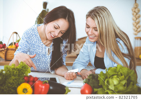 Two women friends choosing the recipe for a delicious meal while sitting at the table in the kitchen. Tablet pc is the best cookbook 82321705