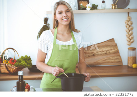 An attractive young blond woman preparing soup by new vegetarian recipe while standing and smiling near the kitchen stove. Cooking and householding concepts 82321772