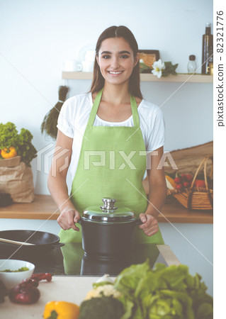An attractive young dark-haired woman preparing soup by new keto recipe while standing and smiling in the kitchen. Cooking and householding concepts 82321776