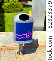 Blue metal trash can with ashtray near the lawn. Urban environmental care concept. Modern stylish waste bin on tiled brick sidewalk as an element of city infrastructure. Dnipro, Ukraine, 2021 82323379
