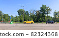 A yellow new car is driving along the road, standing out in the general traffic. A park with green trees, road signs, and a dividing strip with posts In the background. Driving school concept 82324368